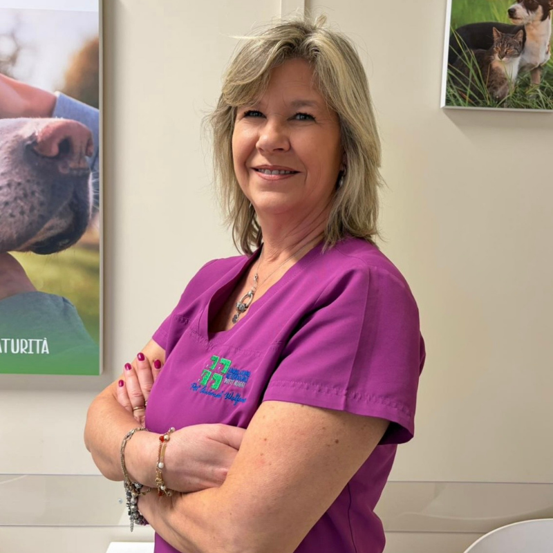 29.1 Primo piano di Cinzia De Alessandri, front office dell'Ambulatorio Veterinario Botti Maggi. Una donna dai capelli biondi con una divisa viola sta sorridendo con le braccia incrociate in una clinica veterinaria.